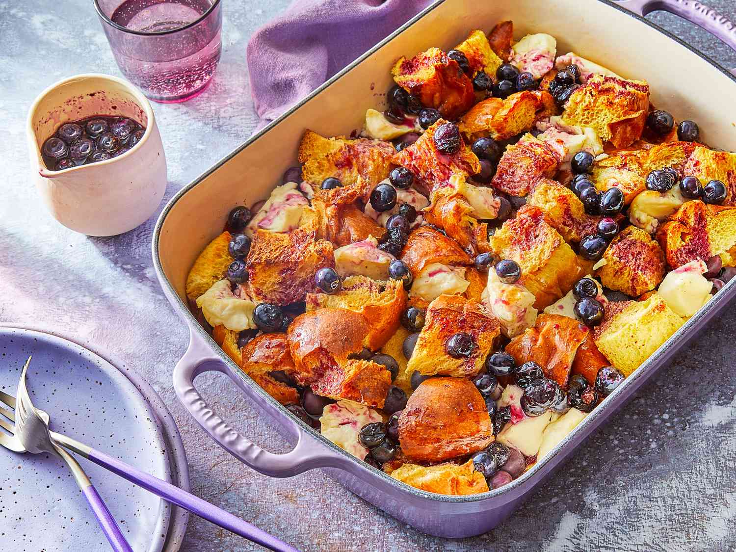 A baked French toast casserole with a crisp golden top, scattered fresh blueberries and raspberries, and a light dusting of powdered sugar