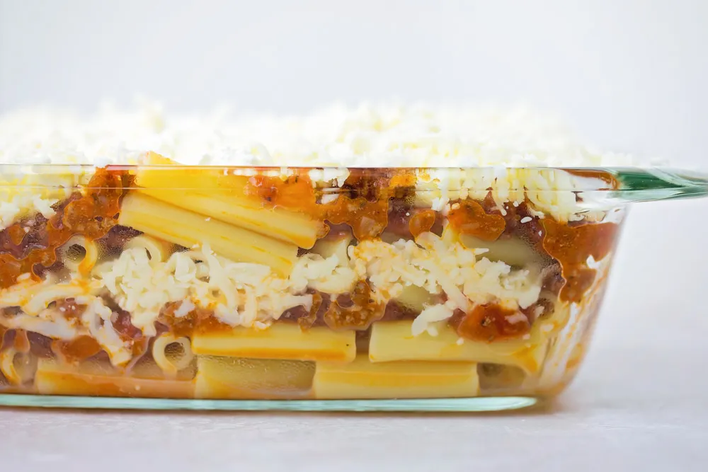 A baking dish being filled with sauced ziti and dollops of ricotta mixture before topping with shredded mozzarella