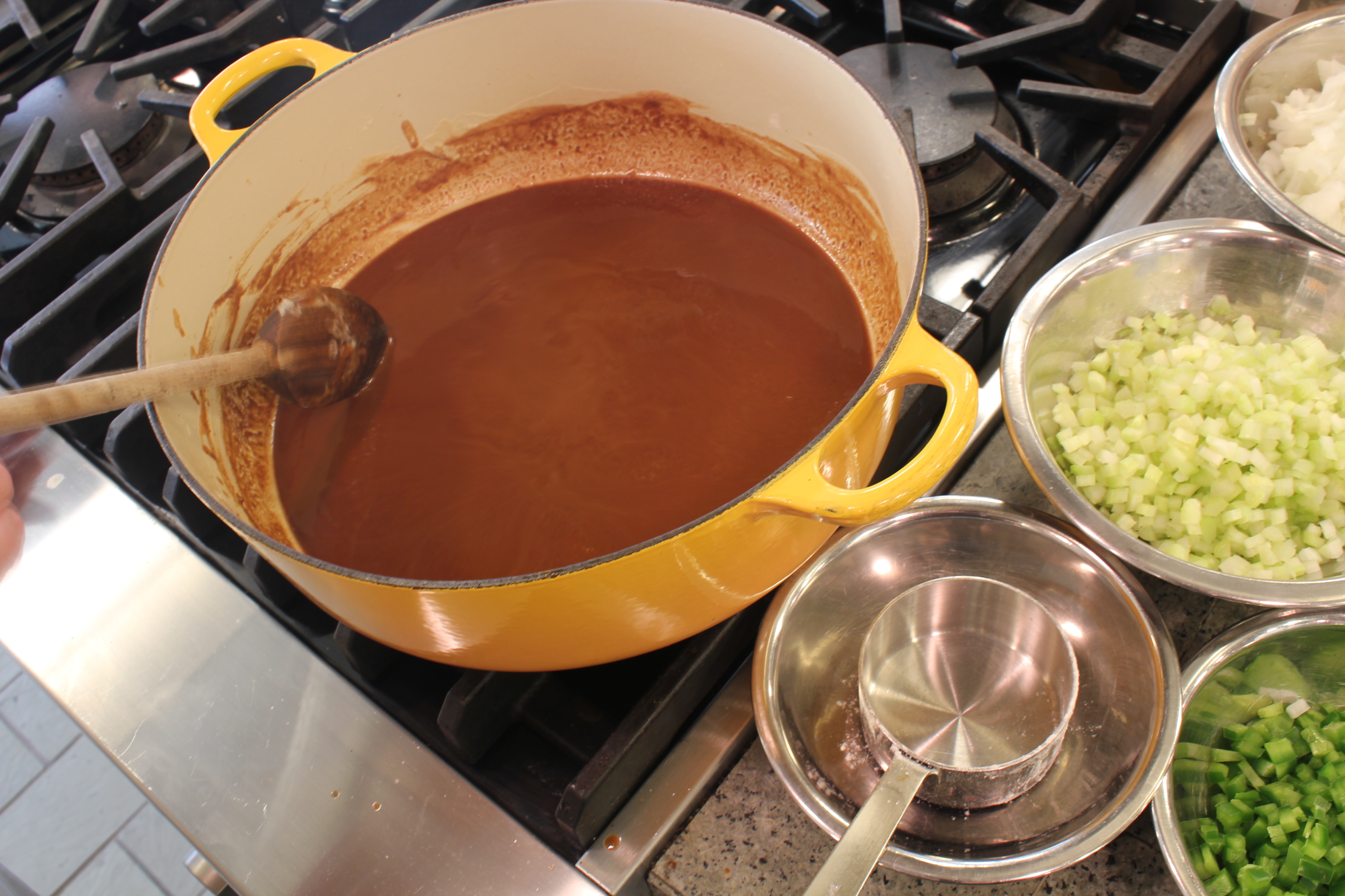 A dark copper-brown roux being stirred with a wooden spoon in a heavy pot on a stovetop, showing a smooth glossy texture