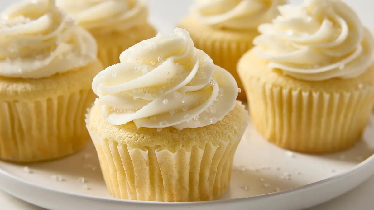A dozen vanilla cupcakes on a baking tray, each topped with a tall swirl of vanilla buttercream frosting in soft, creamy peaks