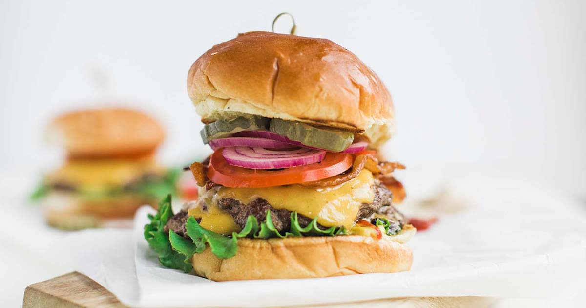 A finished cheeseburger on a plate with shredded lettuce, tomato slices, red onion, and pickles visible in the stack