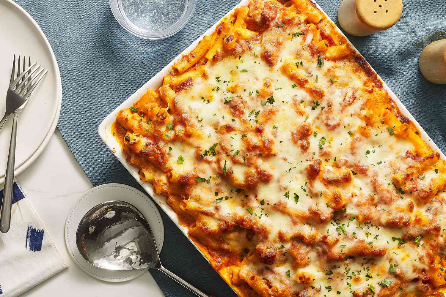 A golden, bubbly baked ziti in a ceramic casserole dish with melted mozzarella and browned edges, a serving spoon tucked into the corner