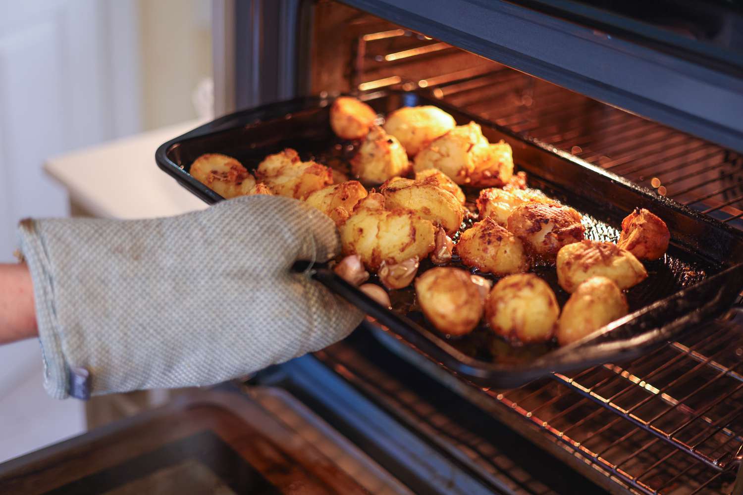 A hand using a metal spatula to flip golden roasted potato chunks on a hot rimmed baking sheet, with visible browned edges and a light sheen of oil