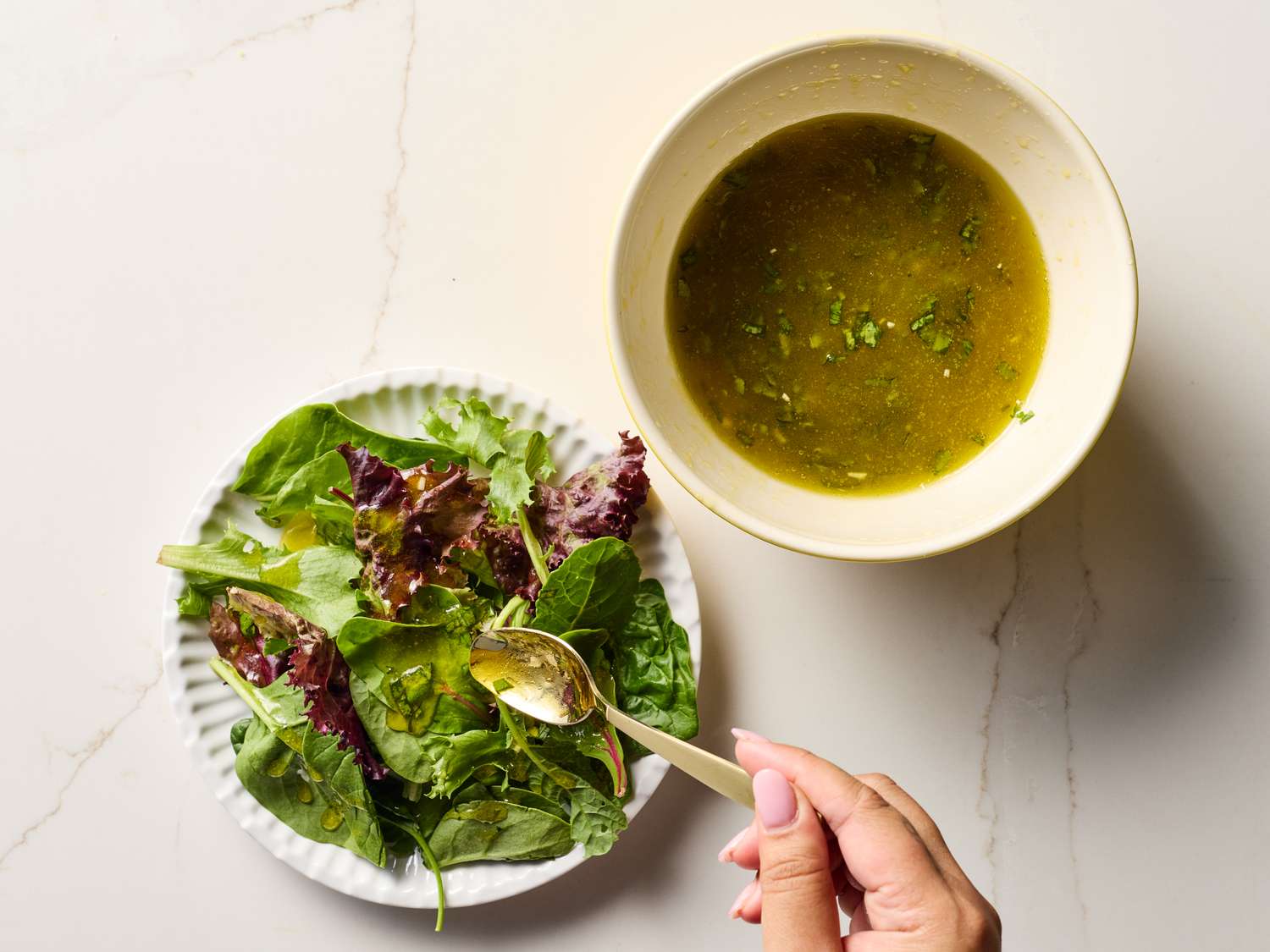 A hand whisking a pale golden vinaigrette in a small ceramic bowl, with olive oil and lemon halves nearby on a kitchen counter