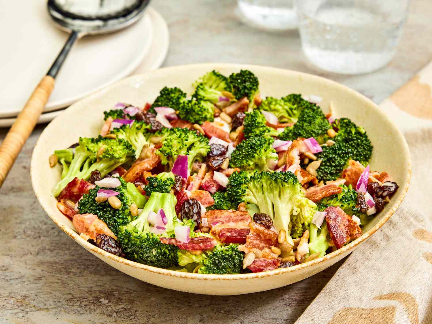A large ceramic bowl filled with broccoli salad with crisp broccoli florets, crumbled bacon, shredded cheddar, and sunflower seeds, set on a wooden table in natural light