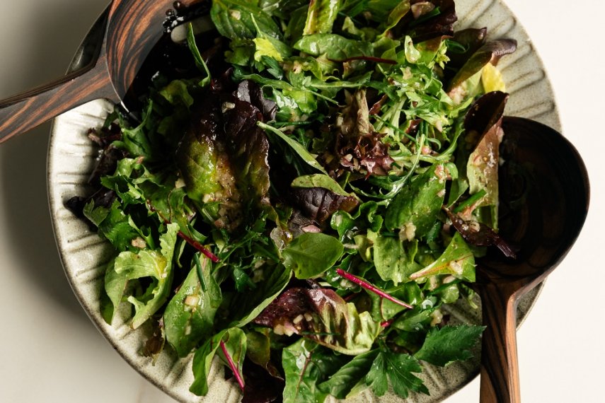 A large glass bowl of mixed salad greens being tossed with a light golden vinaigrette using wooden salad tongs, with droplets clinging to the leaves