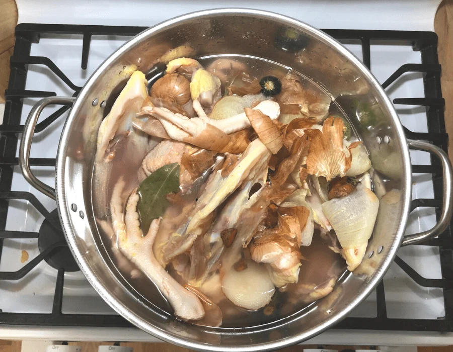 A large stockpot on a stove filled with chicken bones, onion halves, carrots, celery, garlic, and herbs covered with water before simmering