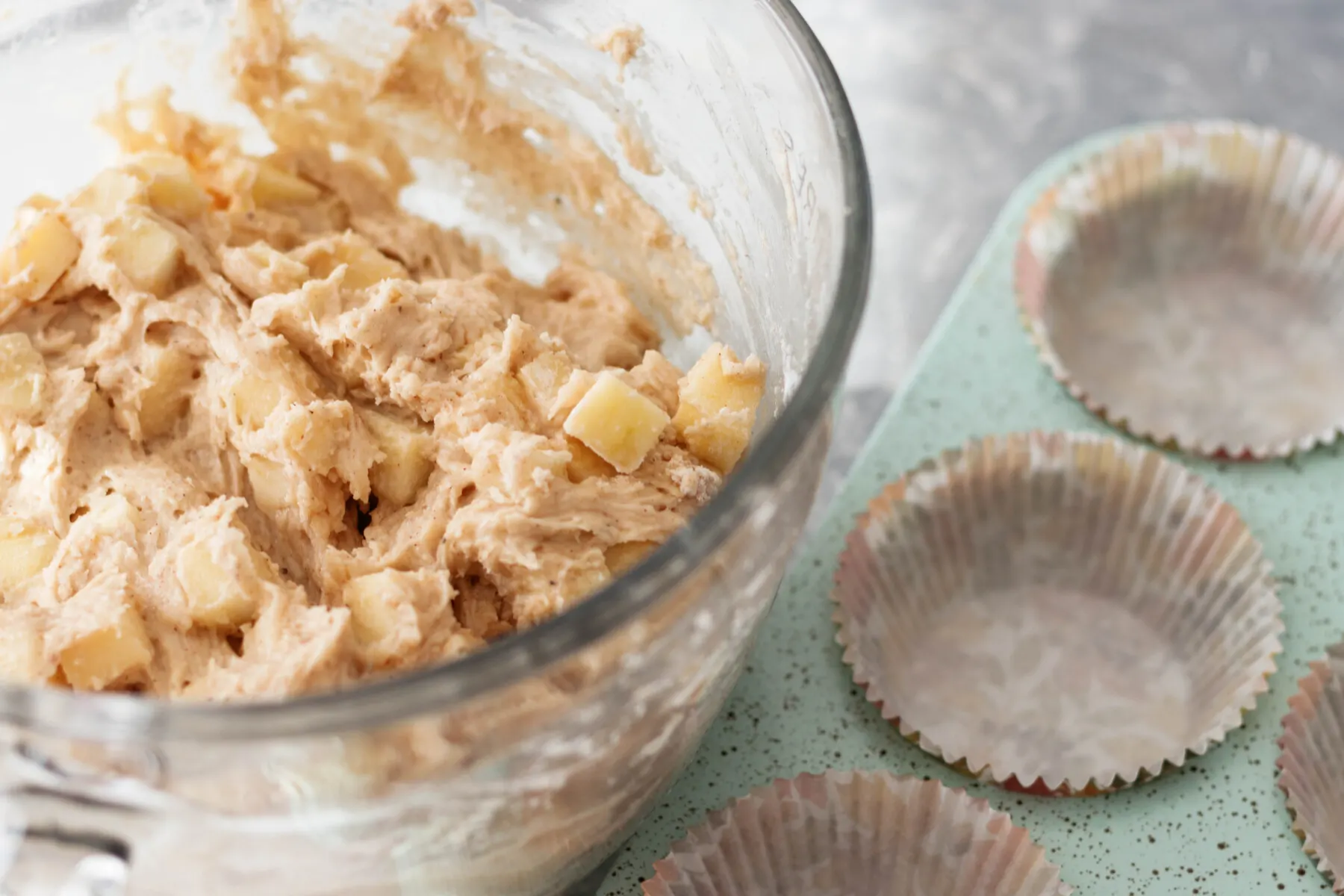 A muffin tin filled with apple-speckled muffin batter, each portion topped with cinnamon sugar before baking