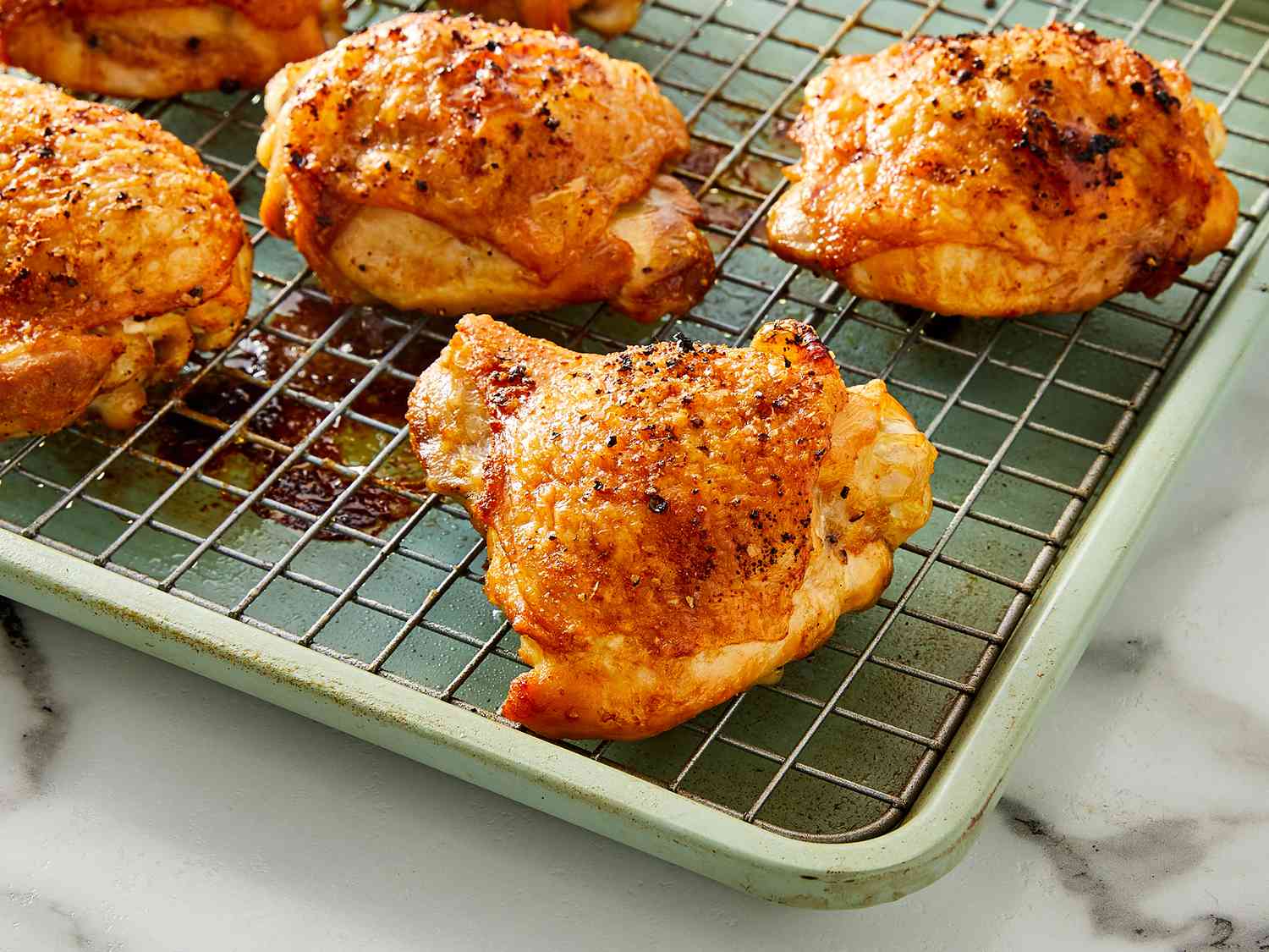 A pan of crispy baked chicken thighs with deeply golden, crackly skin resting on a rimmed baking sheet, with small puddles of rendered fat and browned bits around the chicken