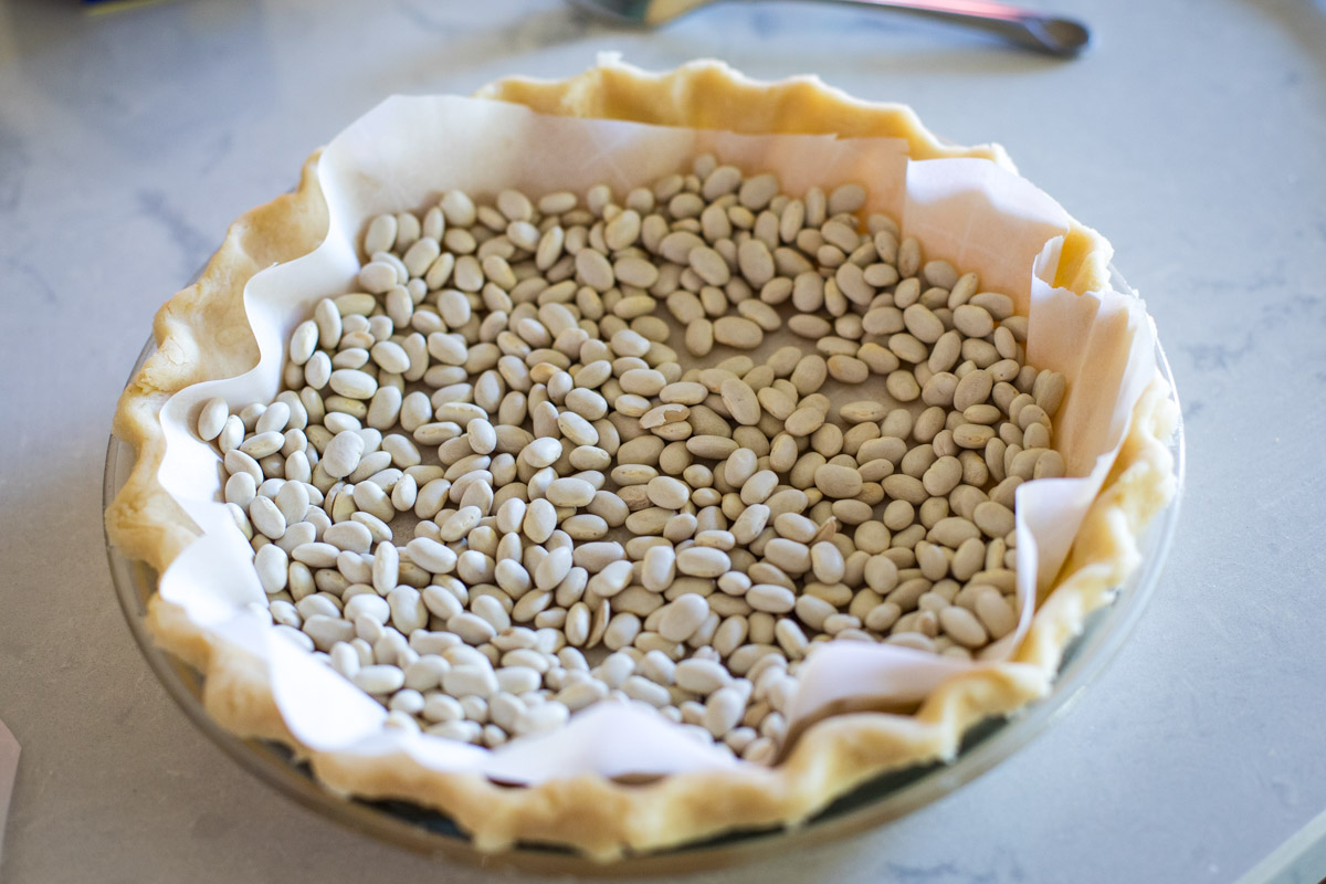 A pie plate lined with raw pie crust and parchment paper filled with dried beans as pie weights, sitting on a baking sheet