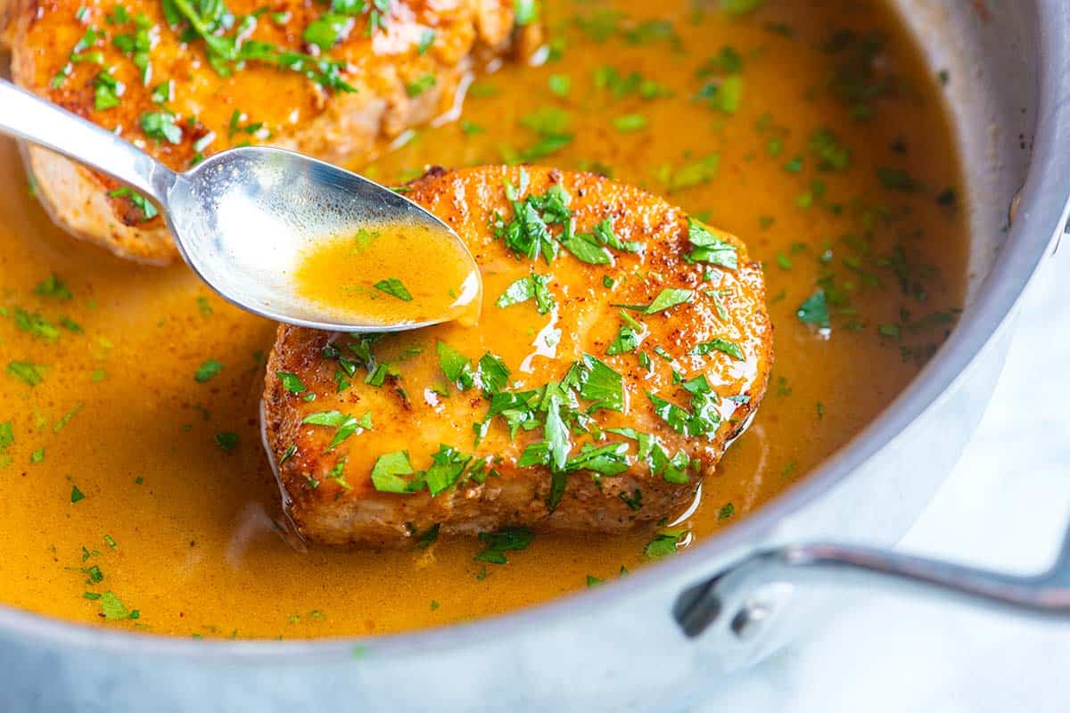 A pork chop resting on a plate beside a skillet filled with glossy pan sauce and browned bits, with a wooden spoon stirring the sauce