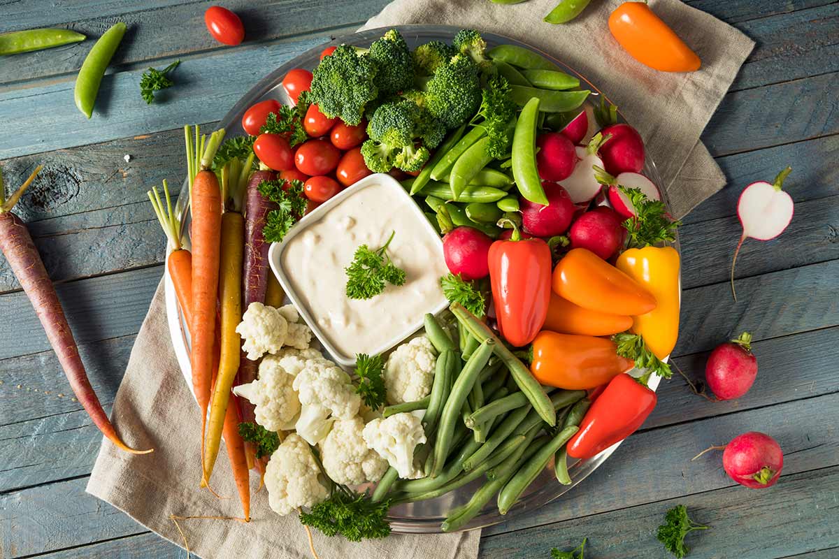 A real photograph of a colorful vegetable tray with carrots, celery, cucumber, and cherry tomatoes arranged around a bowl of ranch dip on a kitchen table