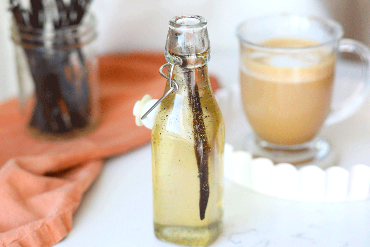 A real photograph of a small glass jar of vanilla syrup with visible vanilla bean specks, a vanilla bean pod resting beside it on a wooden cutting board