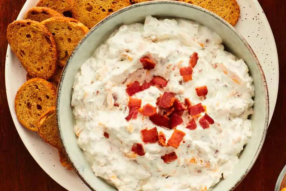 A real photograph of a white ceramic bowl filled with creamy ranch dip, speckled with herbs, set on a wooden board with carrot sticks, celery, and potato chips nearby