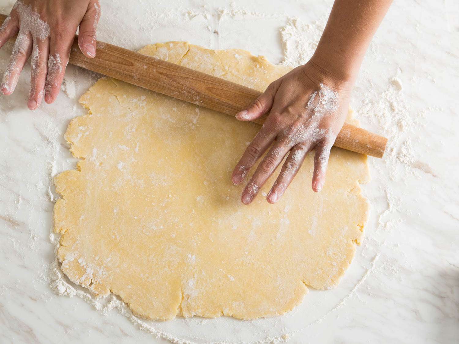 A sheet of pale pie dough being rolled out on a lightly floured countertop with a wooden rolling pin, with visible small butter flecks in the dough