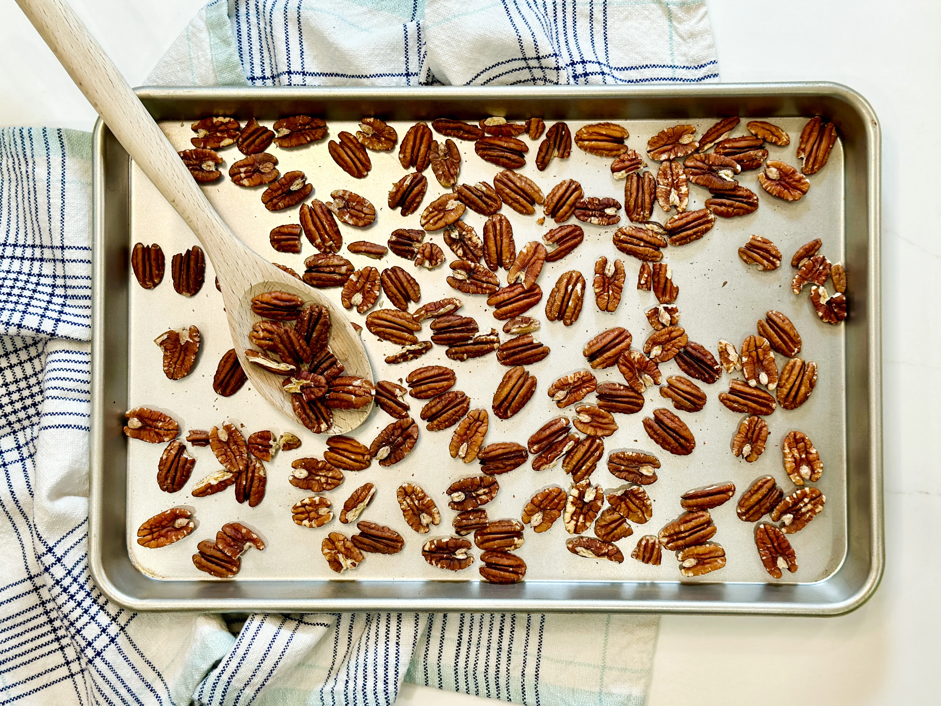 A sheet pan of toasted pecans fresh from the oven, lightly browned and glossy, with a wooden spoon nearby