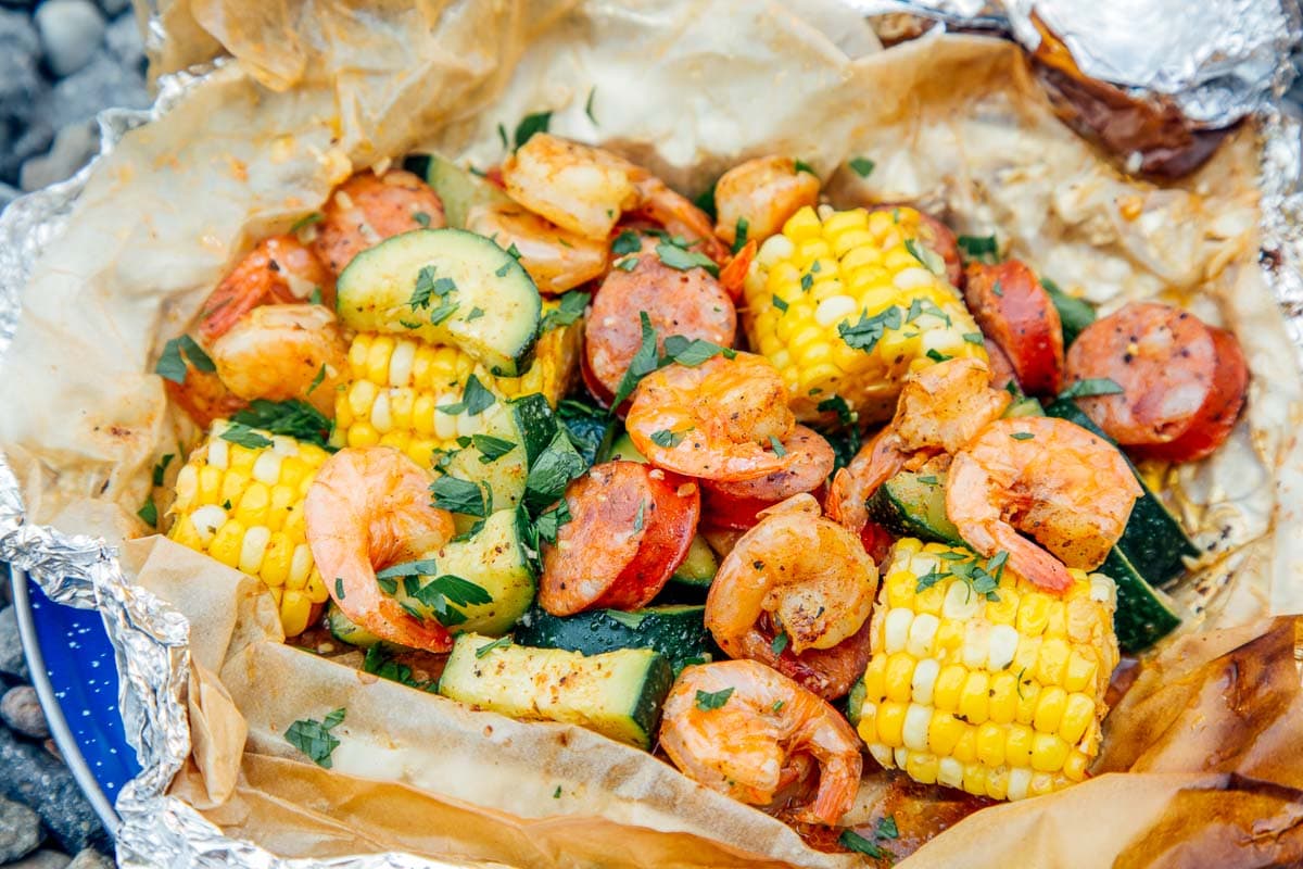 A shrimp boil poured onto parchment paper on a dining table with hands reaching in to serve shrimp, corn, and potatoes
