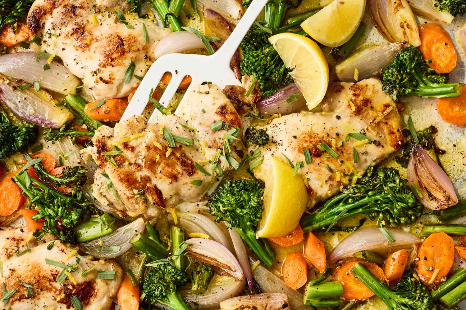 A single sheet pan filled with roasted bite-size chicken pieces, broccoli florets, bell pepper strips, and red onion wedges with browned edges on a parchment-lined pan