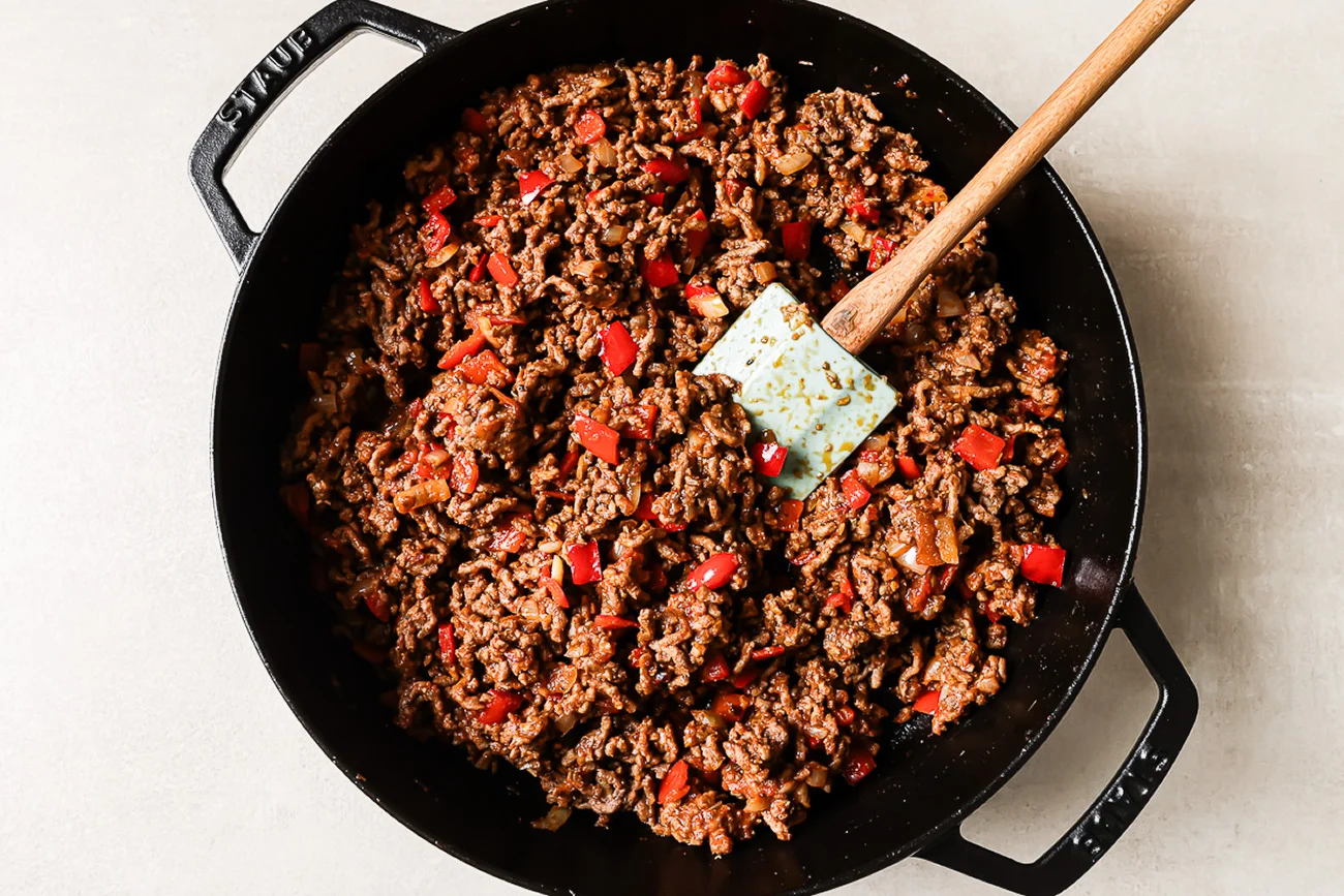 A skillet of thick meat sauce with ground beef simmering in tomato sauce, with a wooden spoon stirring