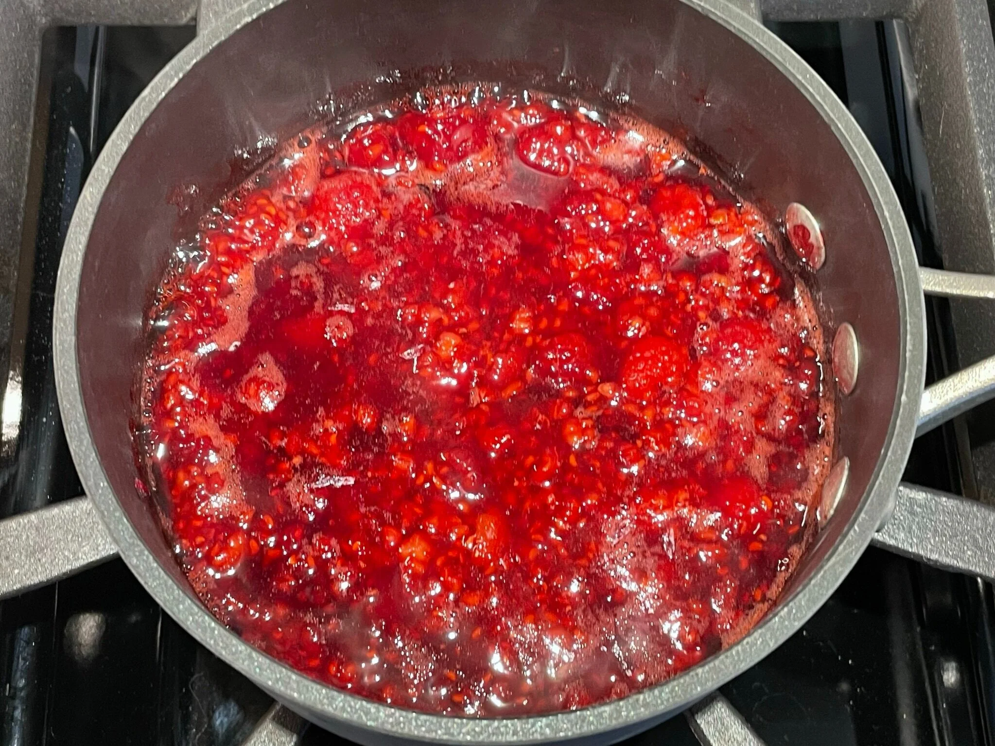 A small saucepan on a stovetop with raspberries breaking down into a glossy sauce, a spoon resting in the pan