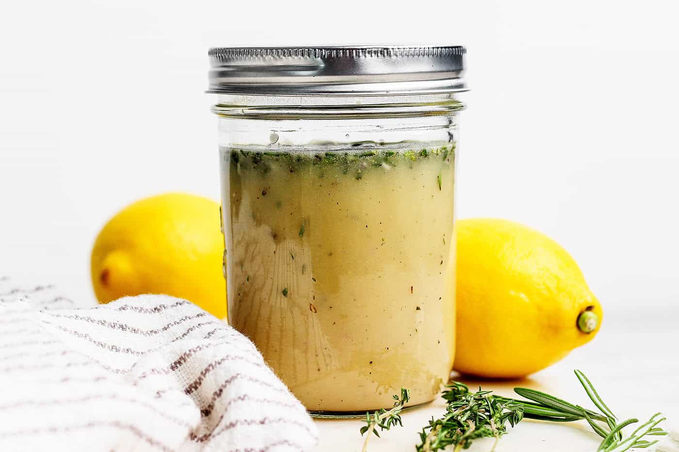 A small sealed glass jar of homemade vinaigrette sitting on a refrigerator shelf next to fresh lemons and a bunch of herbs