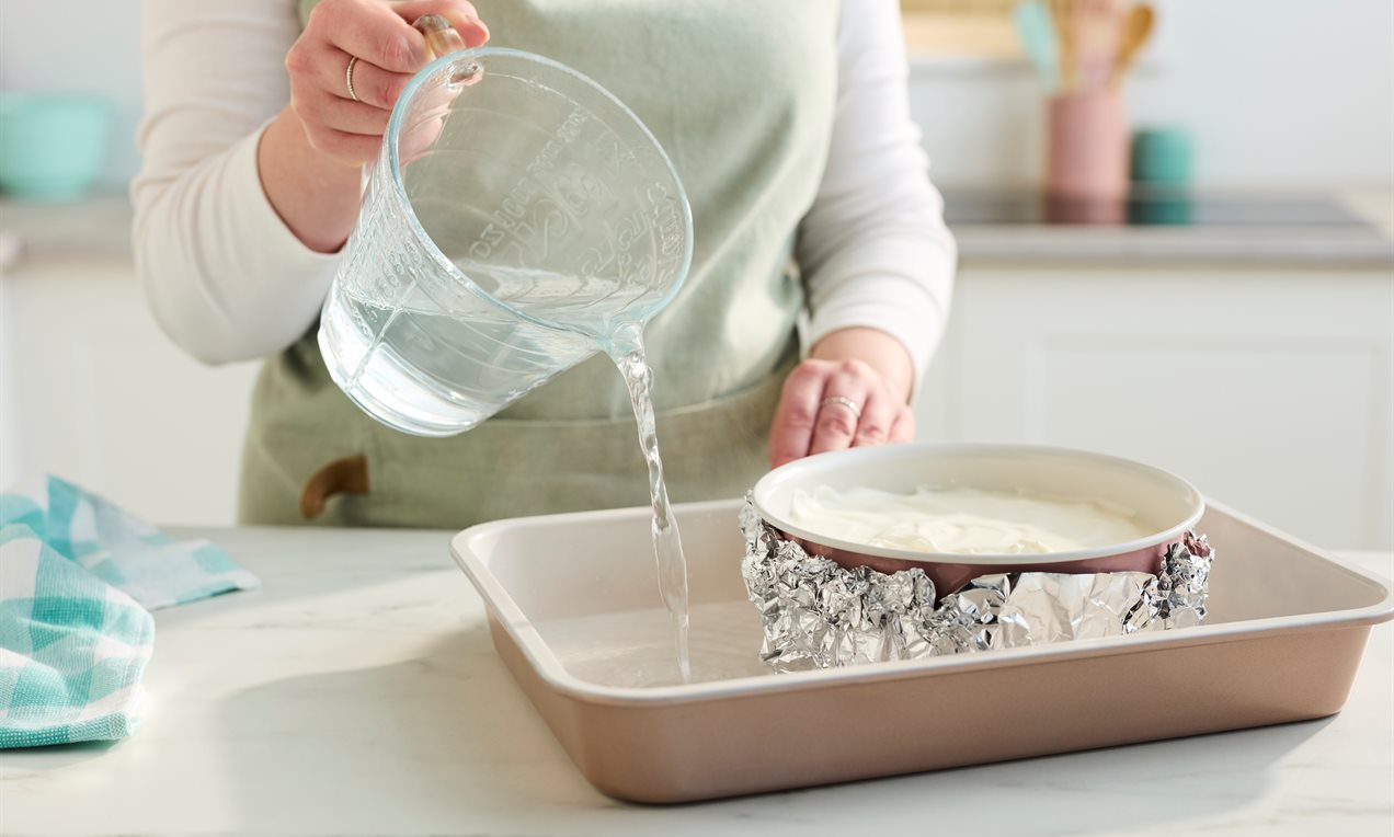 A springform pan wrapped in foil sitting inside a large roasting pan filled with hot water, ready to bake a cheesecake in the oven