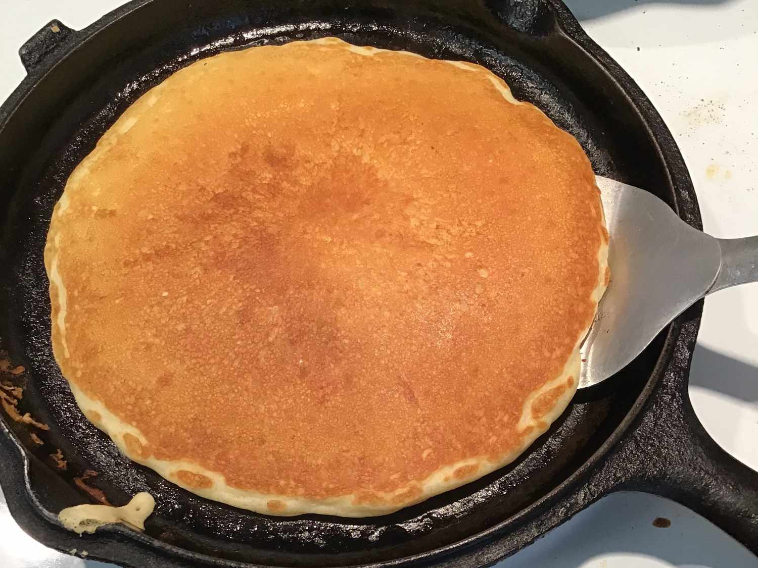 A stack of golden pancakes cooking in a cast iron skillet while a spatula lifts one pancake to flip it