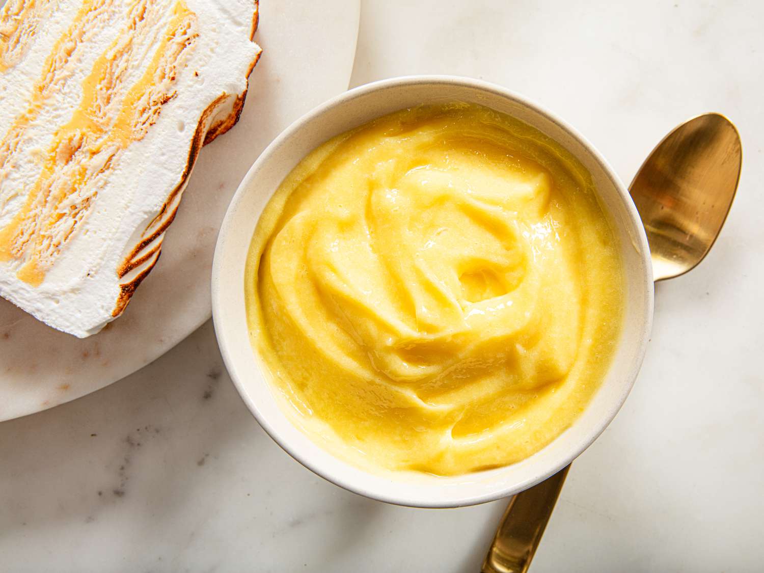 A stainless steel mixing bowl set over a pot of simmering water with a whisk stirring pale yellow lemon curd as it thickens
