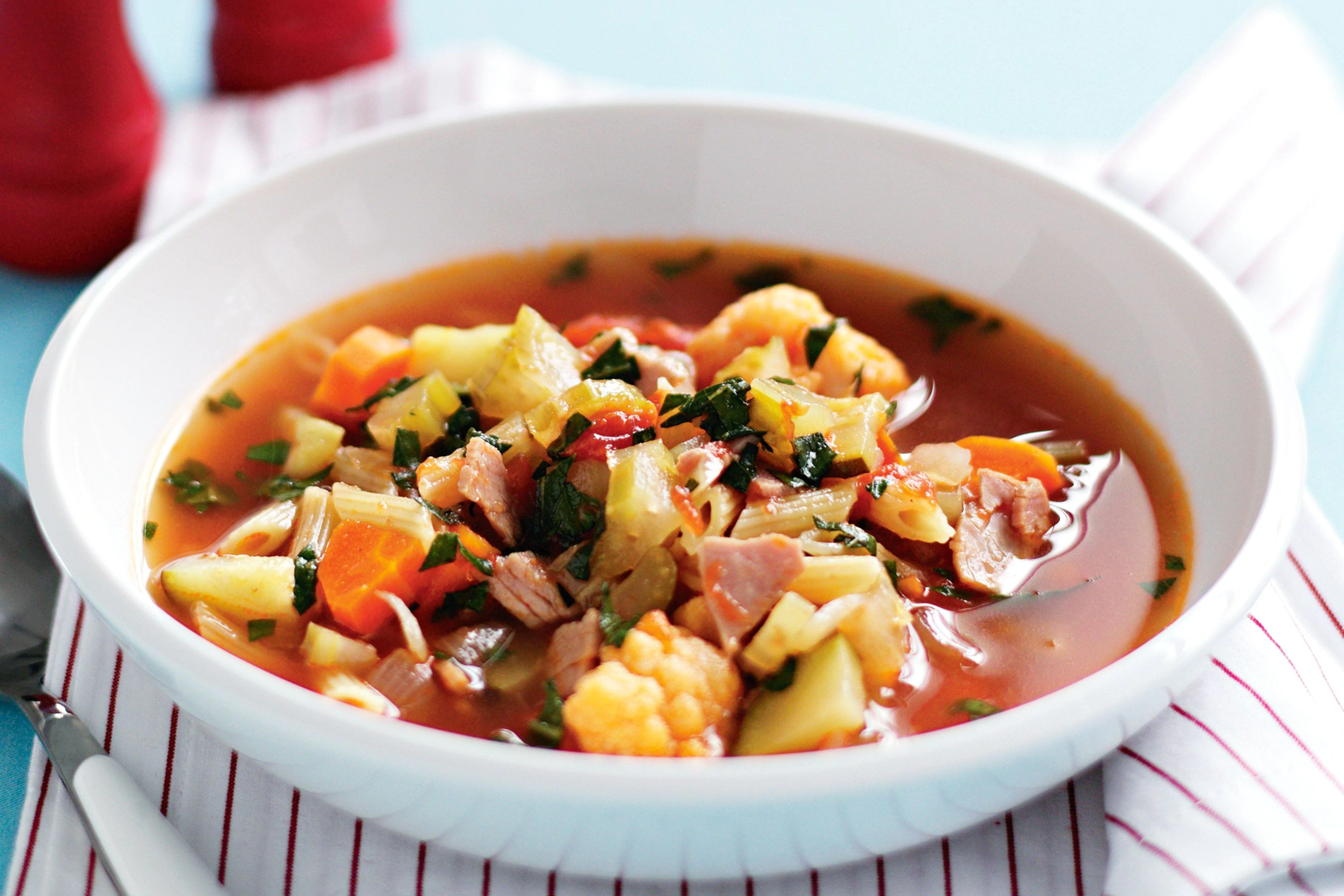 A steaming bowl of hearty vegetable soup with carrots, celery, potatoes, beans, and herbs in a rich tomato broth, photographed on a rustic wooden table with a spoon nearby