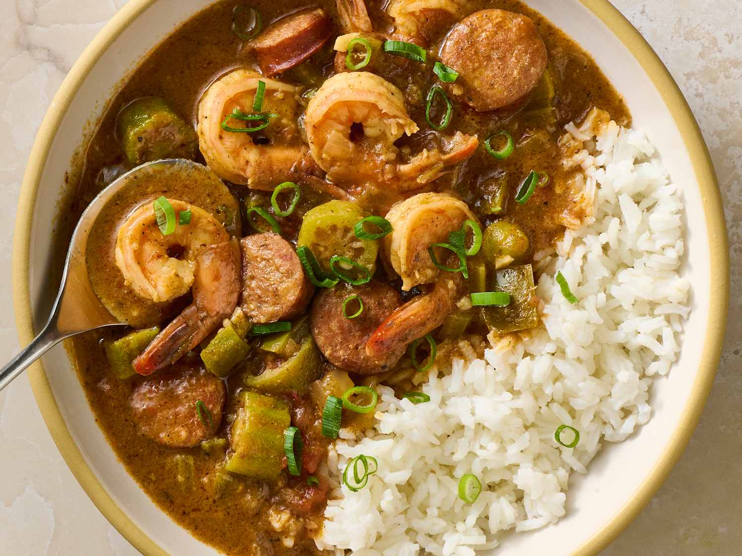 A steaming bowl of shrimp gumbo served over white rice in a wide ceramic bowl, with visible shrimp, okra, and a dark glossy broth on a wooden table