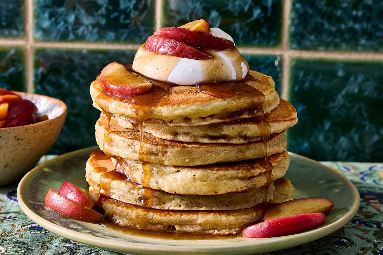 A tall stack of fluffy pancakes on a ceramic plate with butter melting on top and maple syrup pooling around the base on a breakfast table