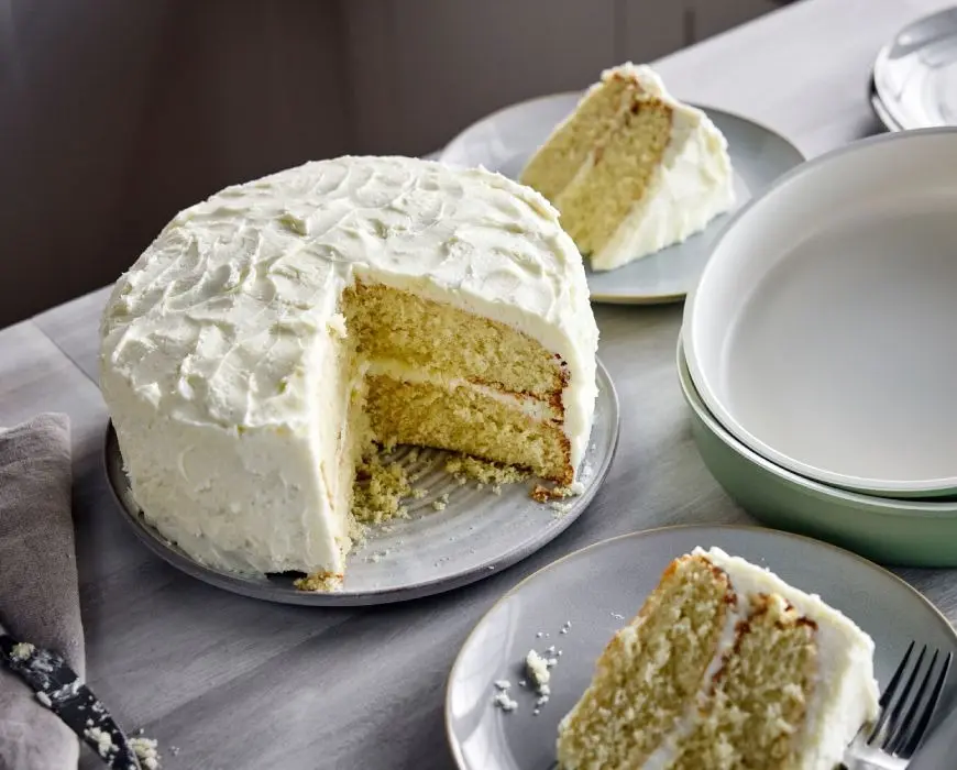A vanilla layer cake on a cake stand being frosted with a spatula, with smooth pale buttercream covering the sides and top in a home kitchen
