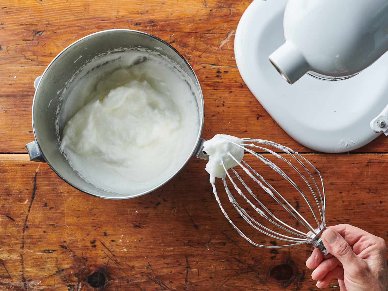 A whisk lifted from a bowl of whipped cream showing soft peaks that droop gently at the tip, with a smooth glossy surface in the bowl