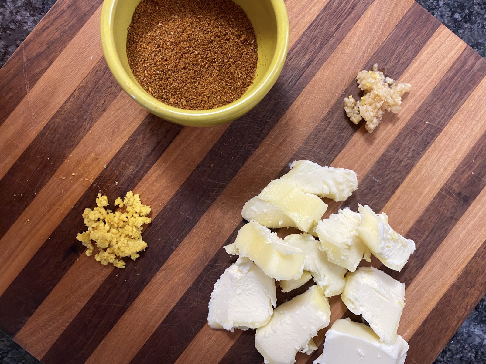 A wooden cutting board with Old Bay seasoning, halved lemons, a stick of butter, garlic cloves, and a knife ready for prep