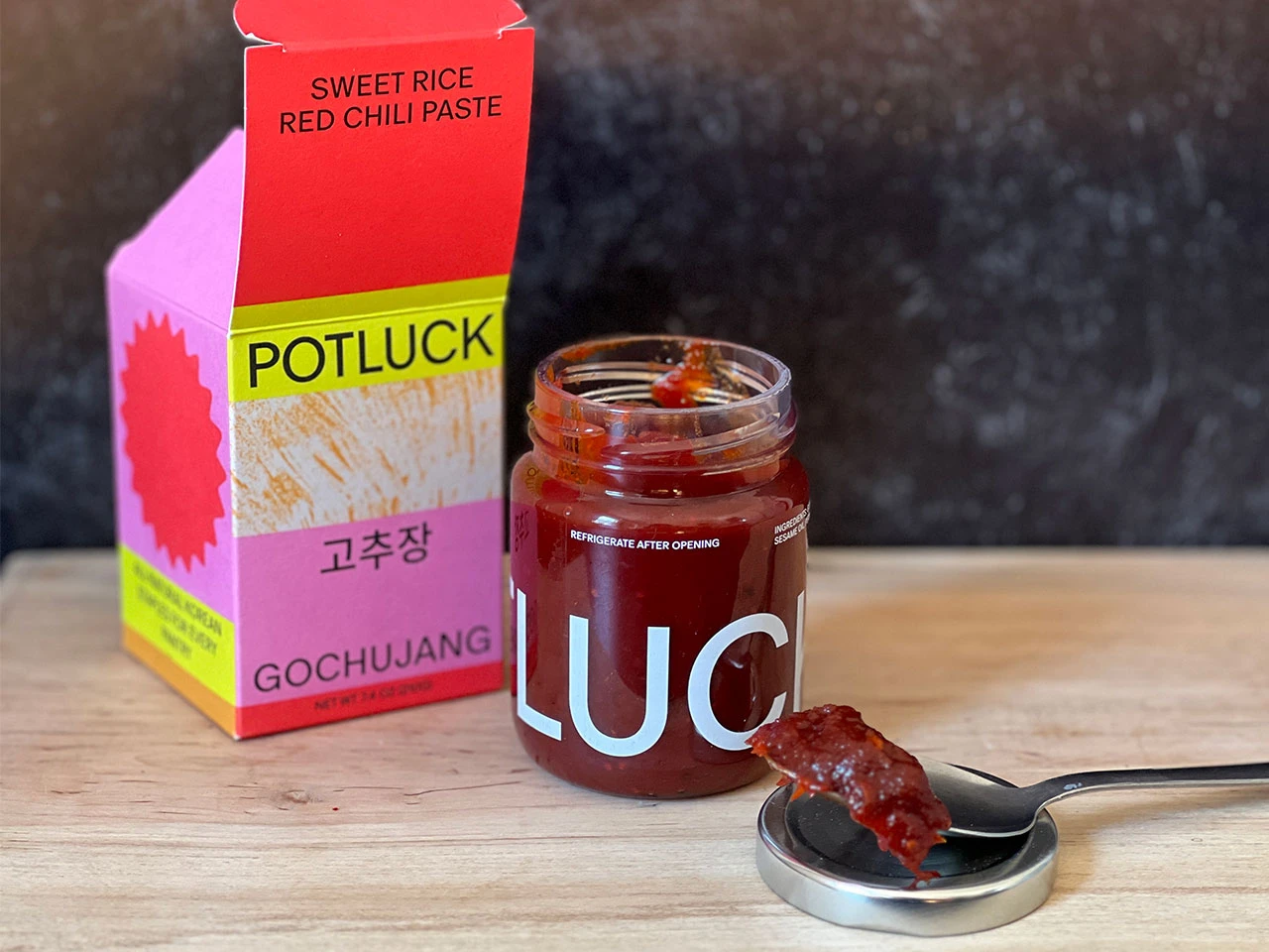 An open jar of gochujang on a wooden countertop with a spoon lifting a thick scoop of deep red paste
