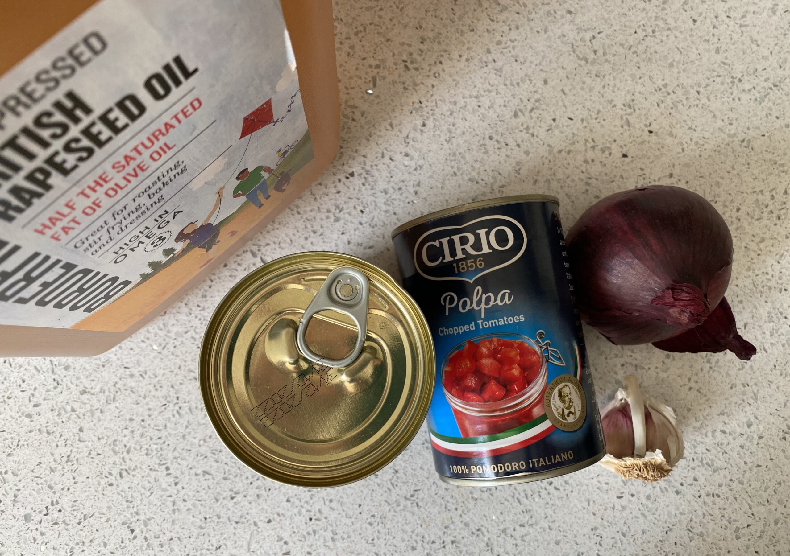 Canned crushed tomatoes, tomato paste, an onion, and garlic cloves arranged on a wooden kitchen counter with measuring spoons