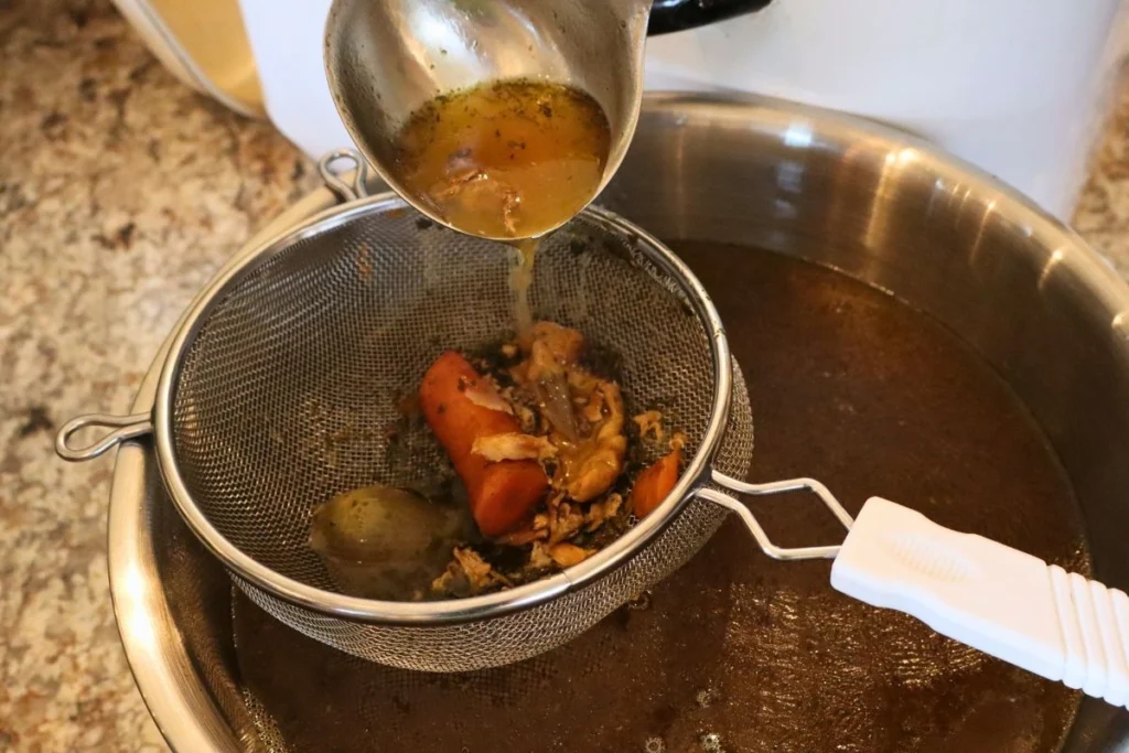 Chicken bone broth being poured through a fine mesh sieve into a large bowl on a kitchen counter, with steam rising