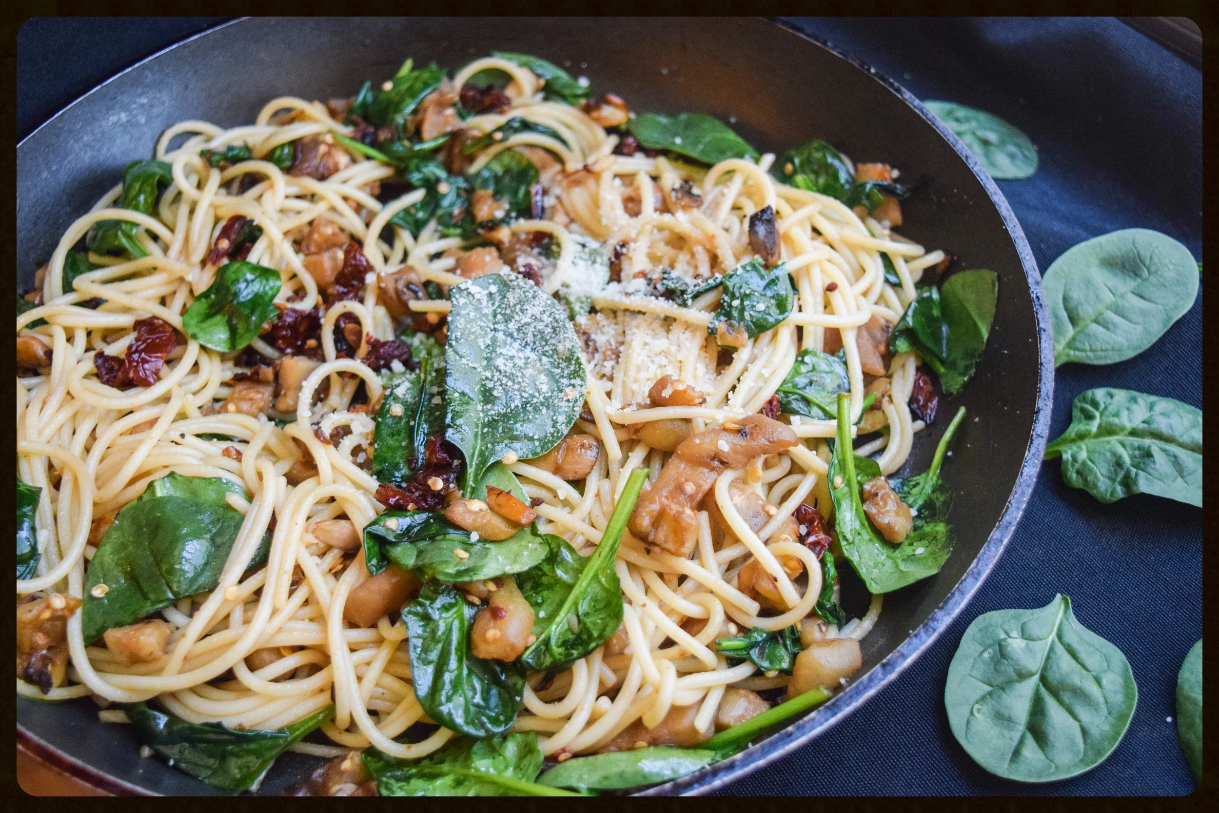 Chopped sun-dried tomatoes, a pile of baby spinach, and minced garlic on a cutting board beside a chef's knife in a home kitchen
