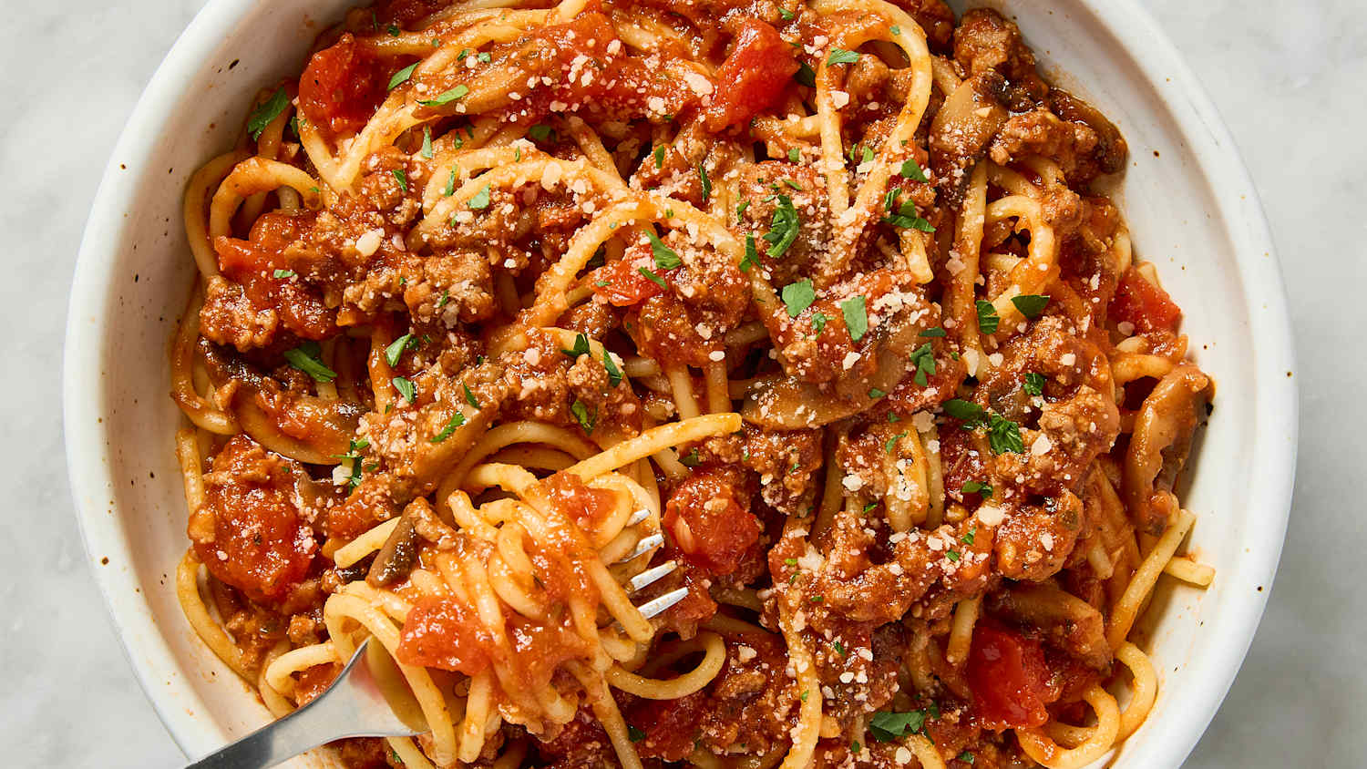 Cooked spaghetti being tossed in a pot with a ladle of meat sauce, steam rising as tongs lift the pasta