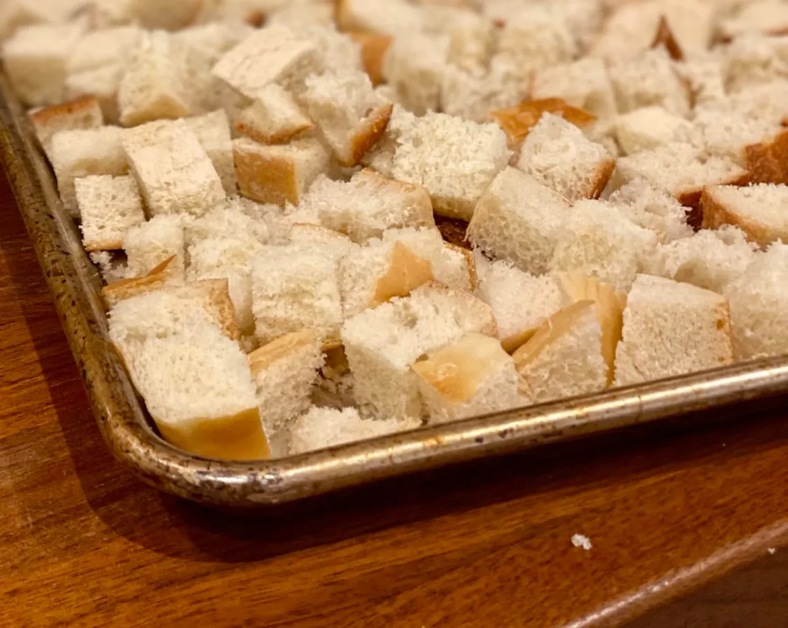Cubed brioche bread spread out on a metal sheet pan, lightly dried and ready to be used for baked French toast