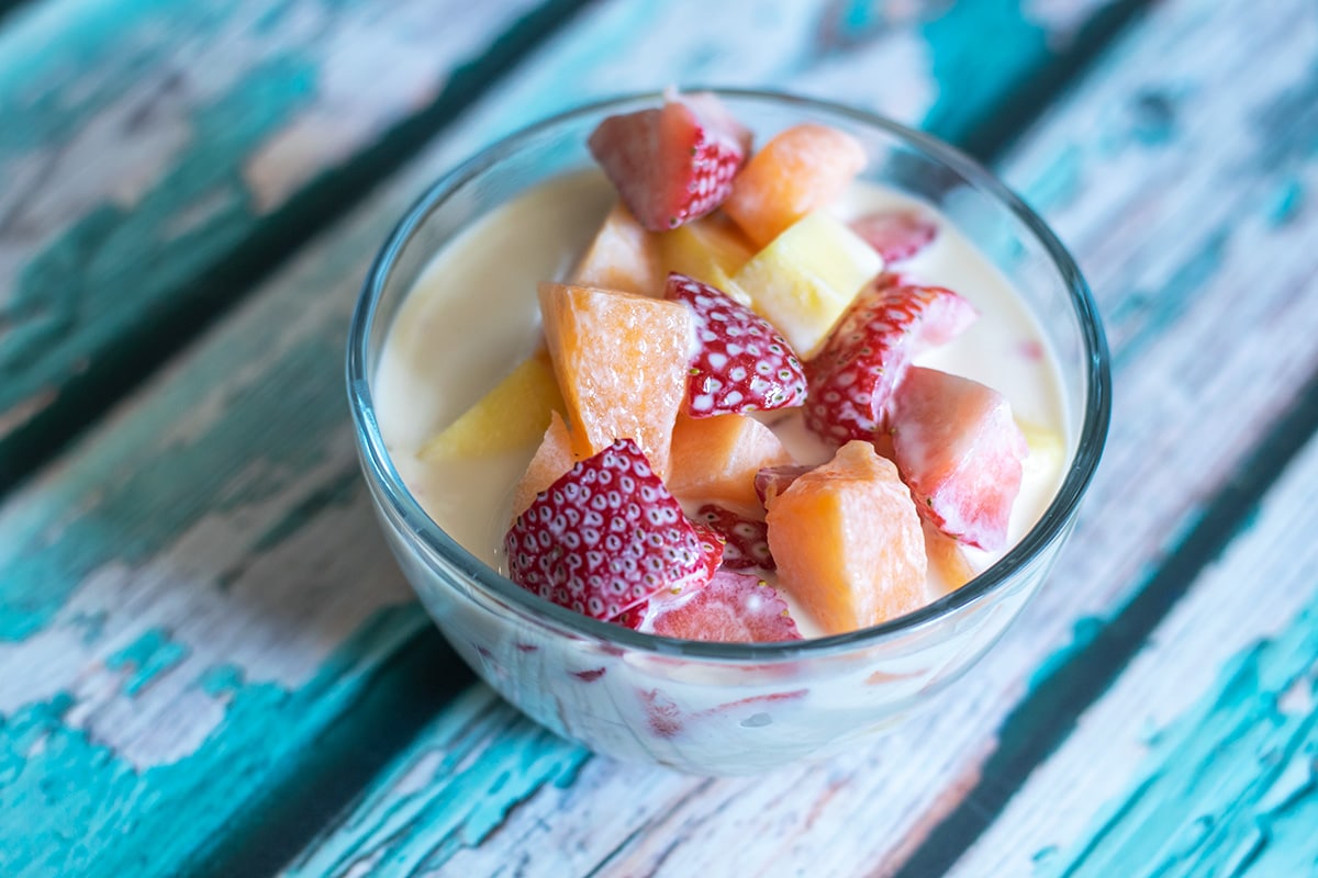 Glass dessert cups filled with mixed fresh berries and kiwi, topped with a pale, glossy condensed milk yogurt cream and a sprinkle of lime zest on a wooden kitchen table