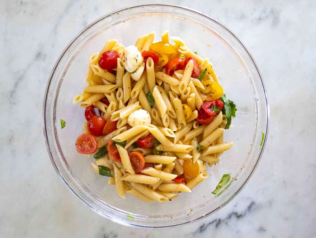 Hands tossing rotini pasta salad with Italian dressing in a large mixing bowl using a wooden spoon, with chopped vegetables visible