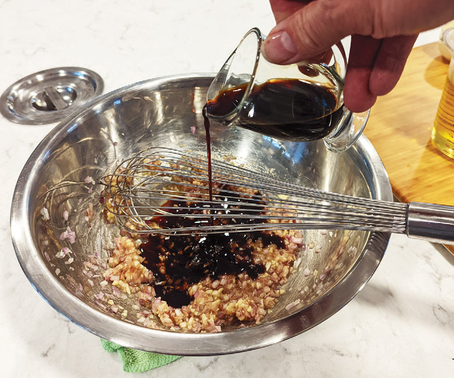 Hands whisking balsamic vinaigrette in a ceramic bowl while olive oil is poured in a thin stream, with black pepper visible on the surface