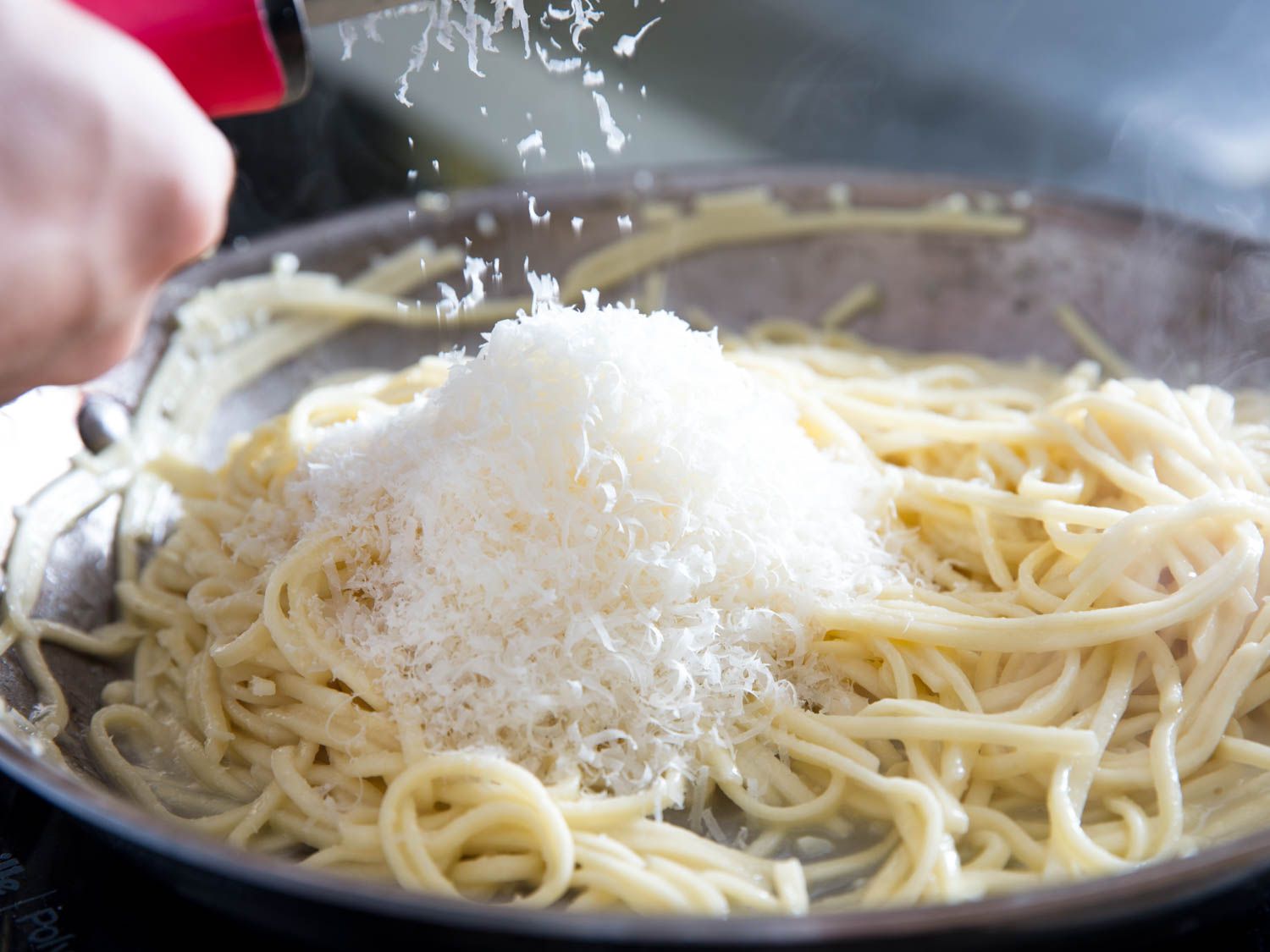 Parmesan being grated on a microplane over a pot of cooked pasta on a stovetop in a home kitchen
