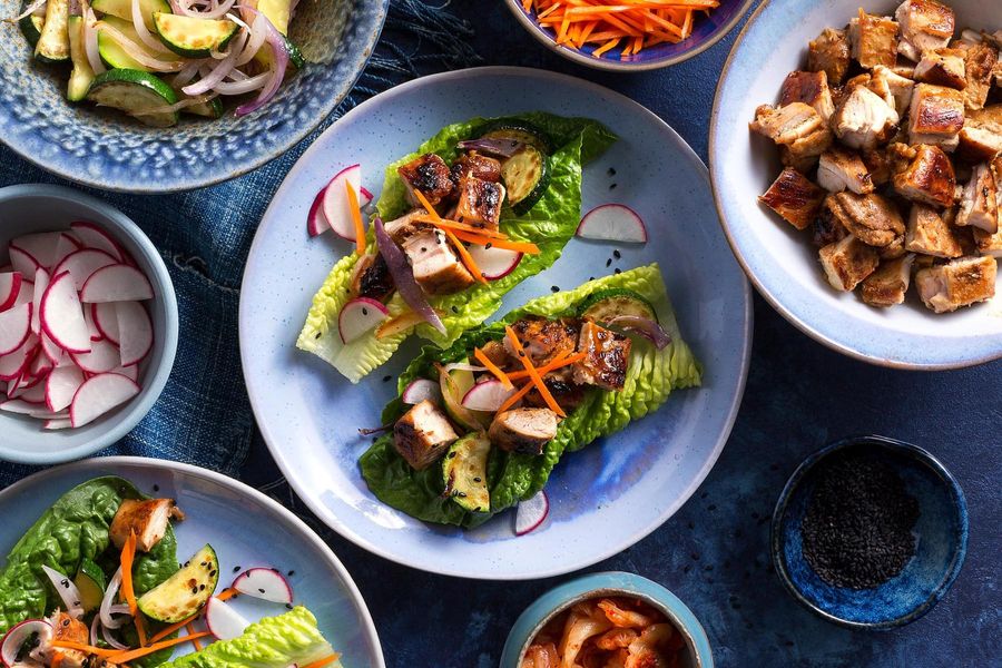 Sliced Korean BBQ chicken tucked into crisp lettuce leaves with white rice, kimchi, and sliced cucumbers on a plate