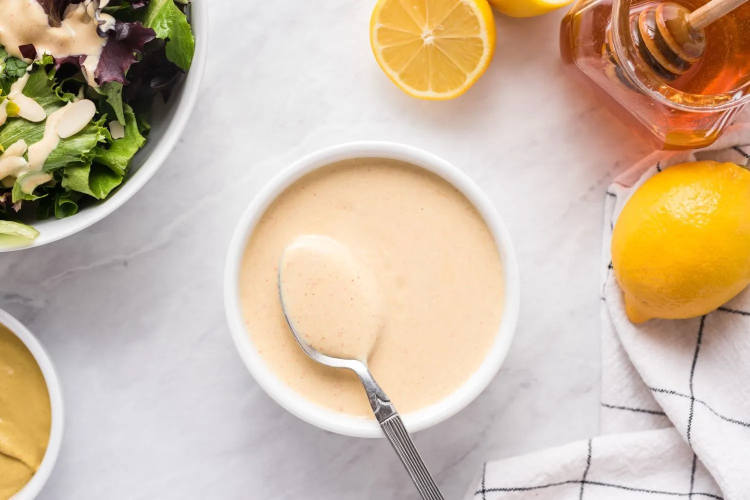 Small bowl of honey mustard sauce being whisked with Dijon mustard and honey, with garlic and a spoon on a wooden countertop