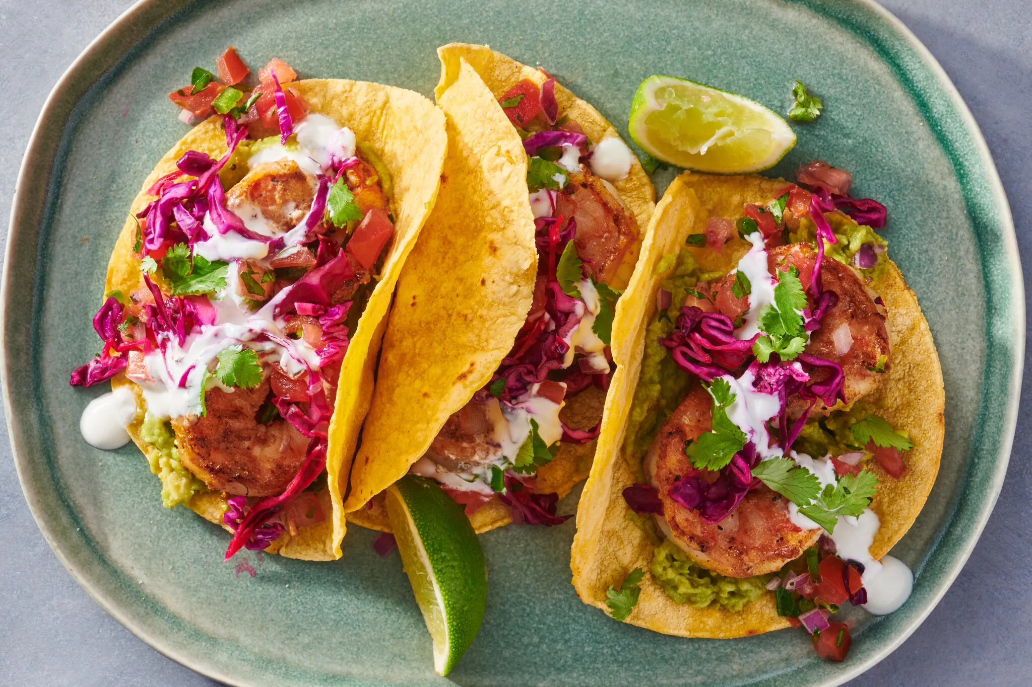 Two shrimp tacos on a plate topped with shredded cabbage, sliced avocado, and chopped cilantro, with lime wedges on the side