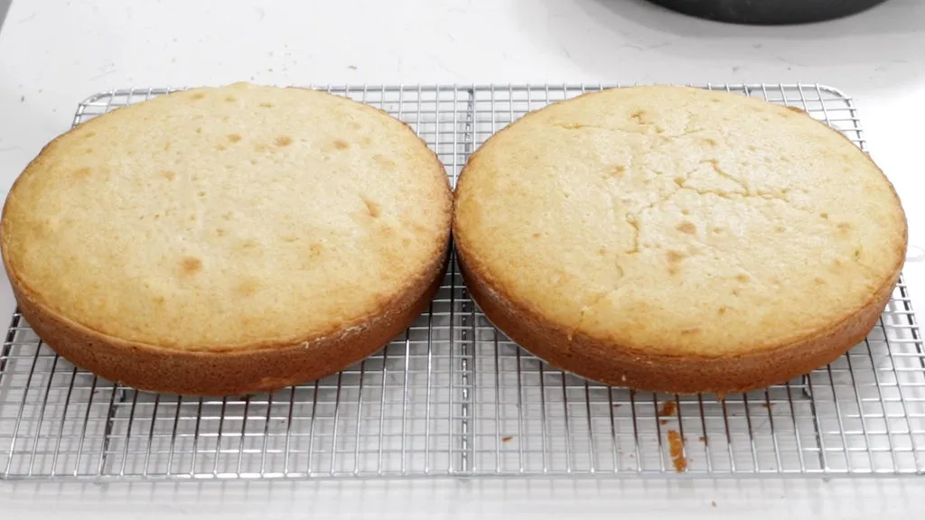 Two vanilla cake layers cooling on wire racks with parchment nearby on a kitchen counter